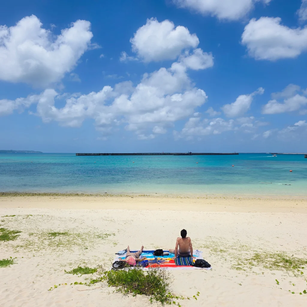 砂浜でレジャーシートを敷いてくつろぐ人と、奥に広がるエメラルドグリーンの海と青い空の風景